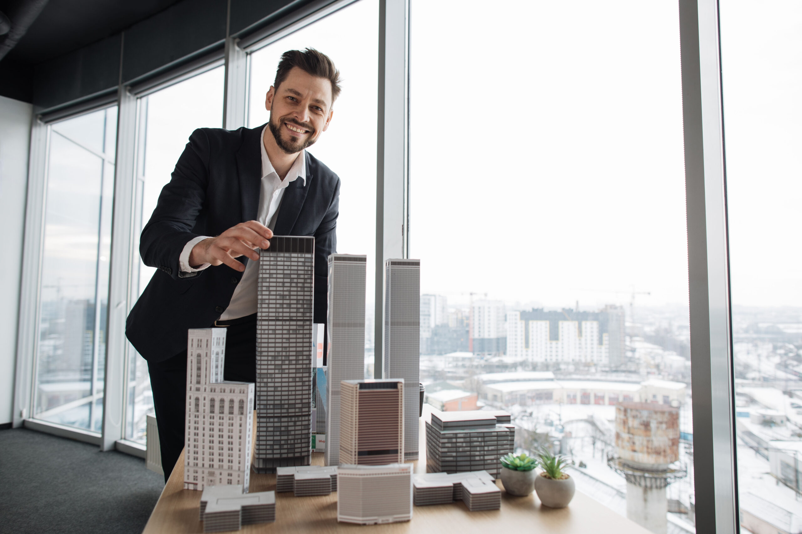 Indoor office portrait of bearded young business man, wearing white shirt and black suit, standing near wooden table with 3D megalopolis model in modern office interior with panoramic city view.